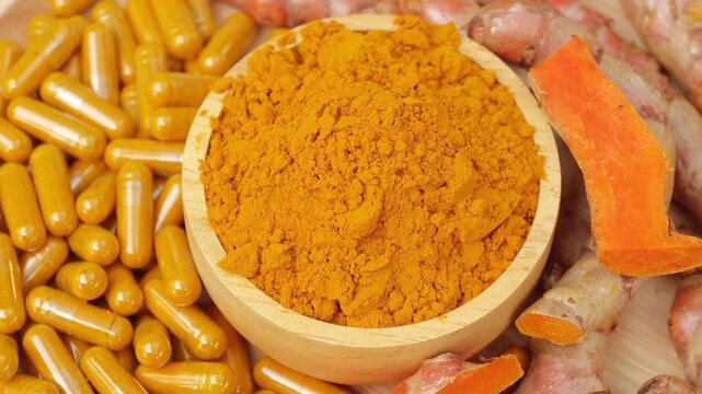 Turmeric powder in wooden bowl surrounded by herbal capsules and fresh curcuma roots