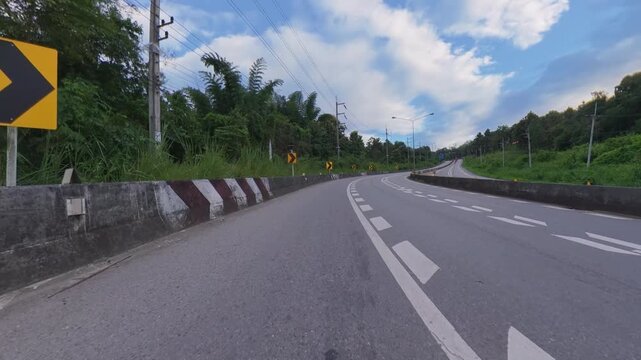 Scenic winding roadway with multiple chevron signs, concrete barriers and lush green vegetation under a blue sky with clouds, showing modern road infrastructure.