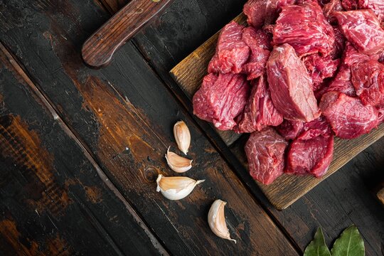 Chopped raw beef chunks arranged on a rustic wooden cutting board beside scattered garlic cloves and fresh bay leaves, set on a dark weathered wooden table under soft overhead light