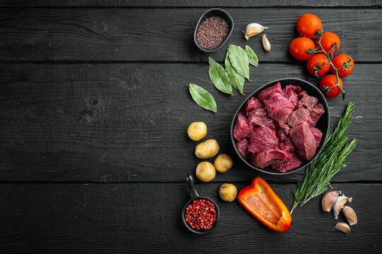 Diced raw stewing beef chunks arranged in a black ceramic bowl beside small potatoes cherry tomatoes on the vine fresh rosemary garlic cloves bay leaves and red peppercorns