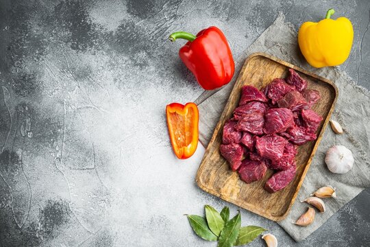 Diced raw stewing beef cubes arranged on a rustic wooden cutting board beside whole yellow and red bell peppers, scattered garlic cloves and fresh bay leaves