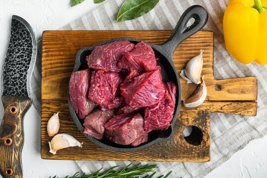 Cast iron skillet filled with diced raw stewing beef chunks resting on a rustic wooden cutting board beside whole garlic cloves fresh rosemary sprigs and yellow bell pepper under bright natural light
