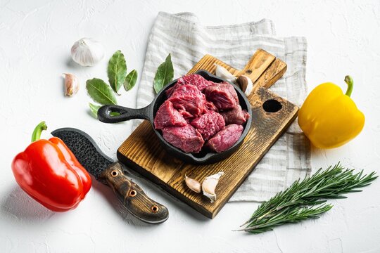 Diced raw beef chunks arranged in a black cast iron skillet resting on a rustic wooden cutting board beside whole red and yellow bell peppers, fresh rosemary sprigs