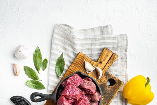 Diced raw stewing beef in a black cast iron skillet resting on a rustic wooden cutting board beside fresh bay leaves and whole garlic cloves, bright yellow bell pepper to the side