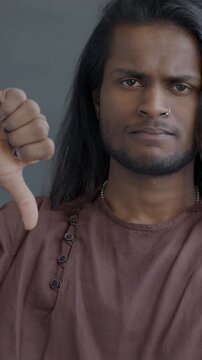 Portrait of unhappy Indian man showing thumbs-down hand gesture shaking head looking at camera on black studio background.