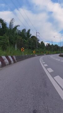Vertical footage of a winding road through lush green forest, featuring multiple chevron signs, concrete barriers, utility poles and a cloudy blue sky.