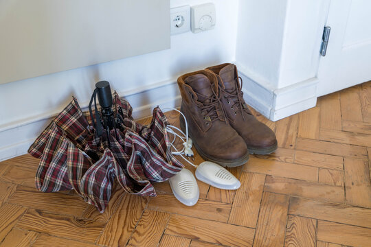 Electric shoe dryers and wet leather boots on a parquet floor beside a folded umbrella in a hallway. Autumn footwear care and drying of damp shoes after rain.