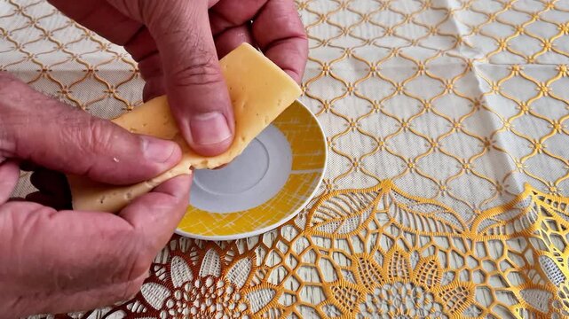 Hands break a folded slice of yellow cheese into smaller pieces over a decorative plate, with an elegant handmade crochet tablecloth creating a cozy traditional food setting