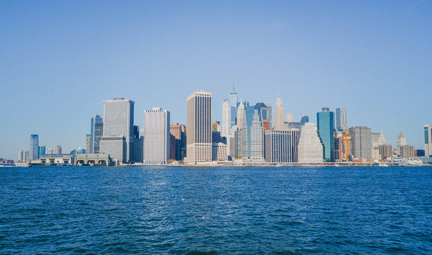 Wide panoramic view of Lower Manhattan from the water, with New York City skyscrapers rising above the waterfront and Hudson River.
