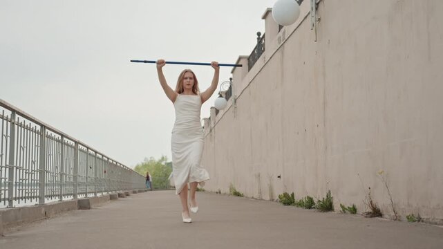 Woman in white dress walks along outdoor pathway beside concrete wall holding and adjusting blue pole overhead. scene features soft natural light with subject moving steadily through calm