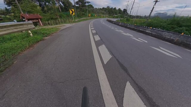 Cyclist POV riding along a winding mountain road with guardrails, chevron signs and concrete barriers, surrounded by lush green forest and distant mountains.