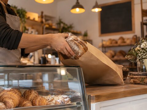 Inside a small bakery counter with a pastry case and hands wrapping a fresh loaf in paper