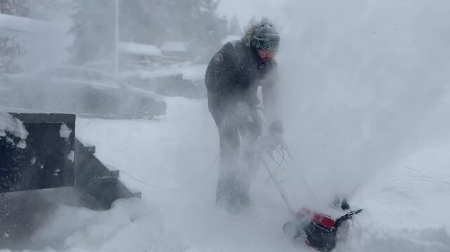 man clearing snow near wooden house in countryside using electric snow blower. Snowing weather conditions. 