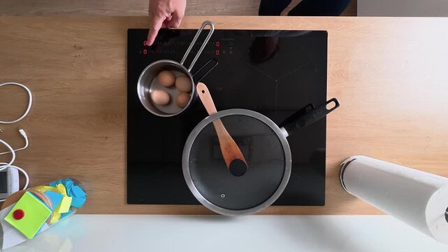 Top-down footage of boiling eggs in a saucepan on a clean induction cooktop, with a covered pan, wooden spoon, and paper towels. Bright, natural light and minimalist decor enhance the modern kitchen s
