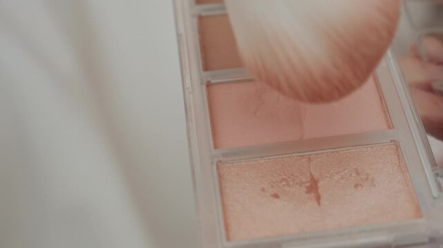 Hand holding makeup brush gently sweeps across various shades of blush and highlighter in open compact revealing texture details. close up view focuses on soft pastel colors like pink