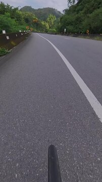 Vertical cyclist POV riding on a winding mountain road through lush green forest, with chevron signs, guardrails and distant mountains along a scenic cycling route.