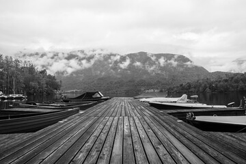 black and white image of wooden dock with boats on calm lake with cloudy mountains in background © Vytenis