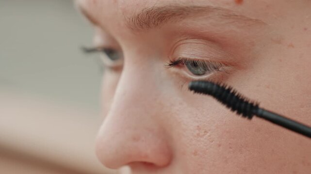 Close up view of woman face as woman applies black mascara to her eyelashes focusing on eye area. scene highlights detailed skin texture and makeup application process with soft lighting