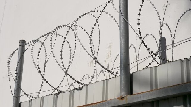 Slow motion close-up of galvanised razor wire coils with sharp barbs mounted on top of a metal security gated fence against an urban industrial wall in Australia perimeter boundary area protection