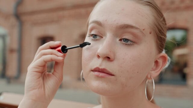 Young woman applies mascara outdoors in front of brick building. Woman holds red and black product focusing on her eye makeup with soft natural light illuminating her skin and jewelry