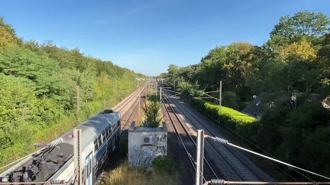 Passenger train traveling along railway tracks through a green wooded landscape, viewed from an elevated position.
