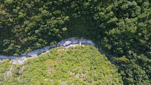 High aerial view of Acheron River winding through dense forest in Greece. Pale rocky banks and Dala Bridge appear below, with sunlit vegetation contrasting against shaded slopes.