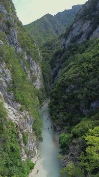 Vertical aerial view of Acheron River gorge in Greece. Visitors wade through pale turquoise water beneath towering rocky cliffs covered in dense green vegetation.
