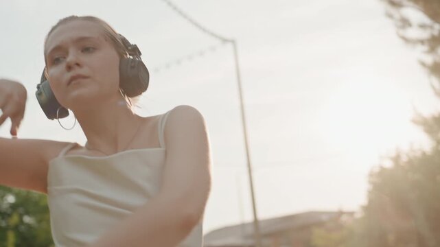 Young woman wearing headphones dances outdoors in white dress moving her arms and body to music under warm sunlight with trees and string lights in background