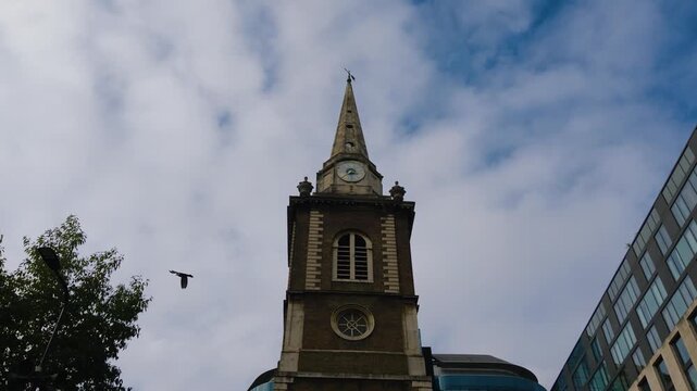 The clock tower and pointed spire of St Botolph&rsquo;s Aldgate rise against clouds beside modern London buildings