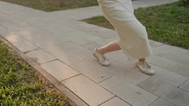 Woman in white dress walks along paved path holding blue exercise bars while wearing headphones. scene is set outdoors with green grass and trees visible under warm natural lighting