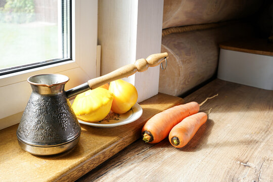 Cozy rustic still life with a copper cezve coffee pot, fresh carrots, and yellow patissons on a wooden windowsill.