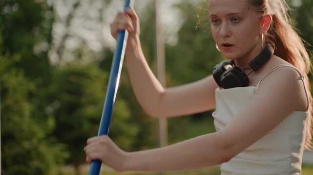 Young woman practices pole vaulting outdoors holding blue bar in various positions. Woman wears headphones around her neck and athletic tank top against green park background with trees