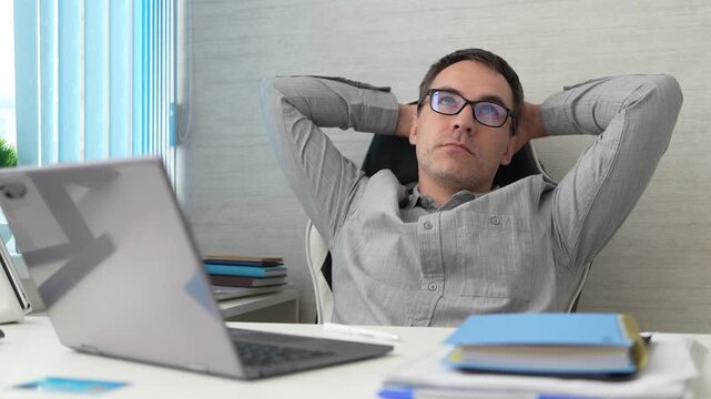 Office worker leaning back in his chair with his hands behind his head, taking a break and looking away from the screen. Rest, relaxation, and completing a task.