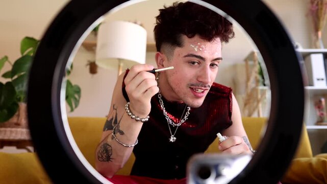 Young non binary person with tattoos and alternative style applying face gems while recording a beauty tutorial video for social media with a ring light and smartphone at home
