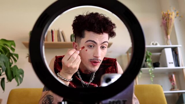 Non binary influencer recording a makeup tutorial for social media, applying red lipstick hearts on their cheeks while vlogging from home with a ring light and smartphone on a tripod