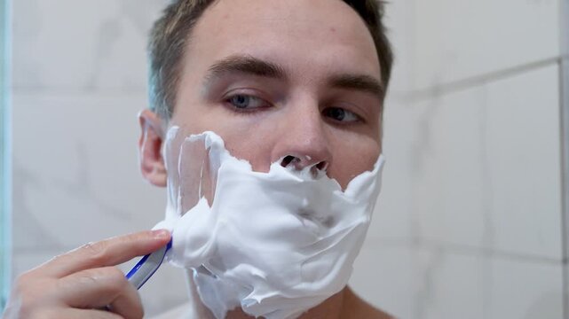 Man applying shaving foam and shaving his face with a razor in bathroom. Grooming routine, personal hygiene, and self care.