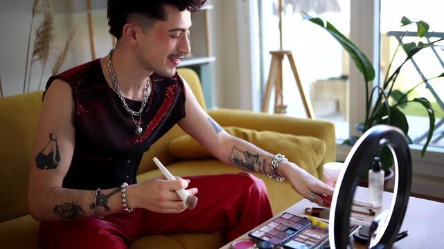 Young non binary influencer with tattoos applying makeup with a brush, filming a beauty tutorial with a smartphone and a ring light for a social media video blog in their living room