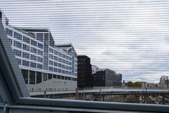 Contemporary buildings seen beyond an elevated urban structure, with layered glass facades and geometric architecture under an overcast sky