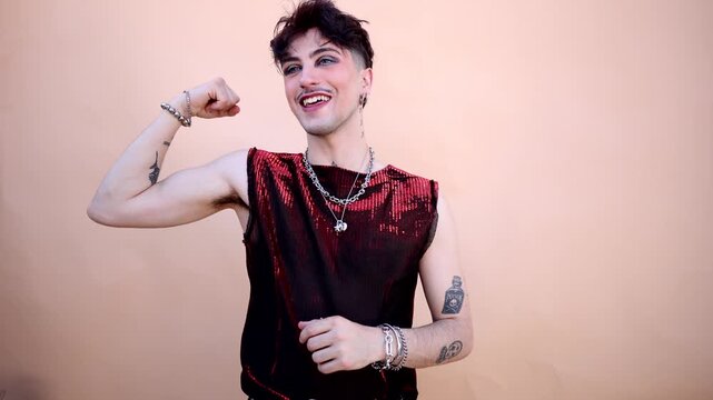Confident non binary person with tattoos, makeup, and mustache smiling while flexing biceps muscles, embodying queer strength and alternative beauty in a modern portrait on a neutral background