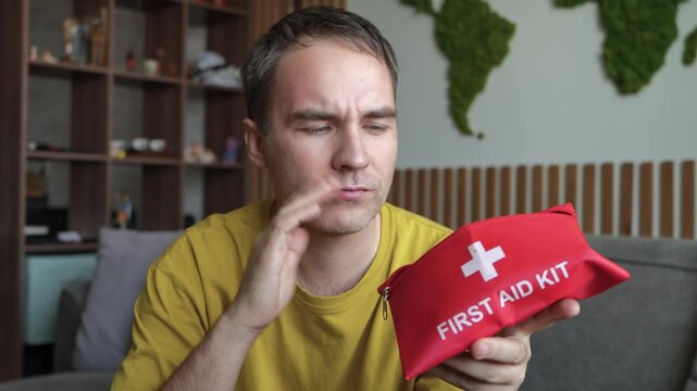 Man searching for painkillers in a red first aid kit, suffering from a severe headache. Migraine, head pain, and urgent medical treatment.