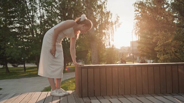 Woman in white dress interacts with her phone and wooden bench beside her bending over to reach out. Woman holds object near waist level while standing on grass next to paved path surrounded