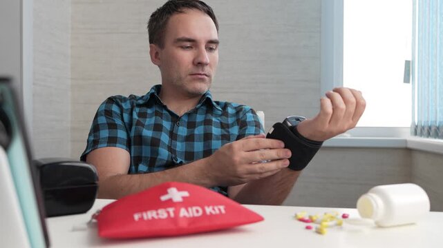 Man measuring his blood pressure at a table with a first aid kit nearby. Hypertension, hypotension, and cardiovascular health monitoring.