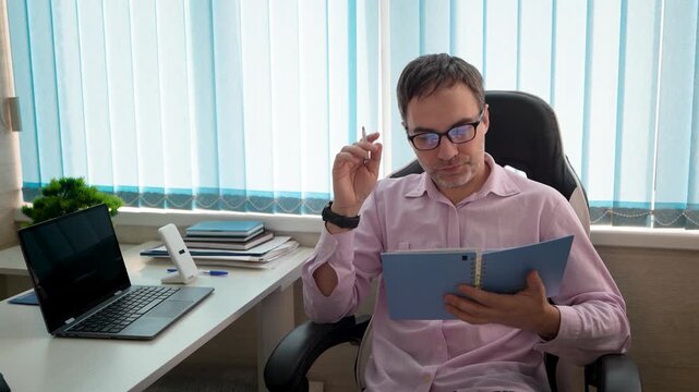 Focused businessman reading notebook in office while reviewing information, studying plans, and preparing for business tasks in a workspace.