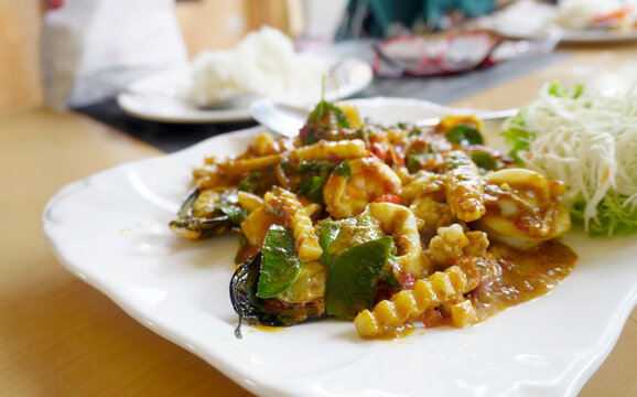 elicious plate of spicy Thai stir-fried seafood with squid, mussels, and herbs on a white plate, served on a table in a restaurant.