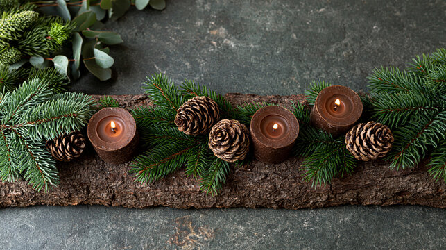 Lit brown candles, pine cones, and fresh green fir branches arranging on a textured wooden log, symbolizing christmas, winter holidays, and advent season