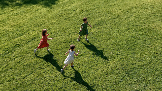 Three happy children running and playing freely on a vibrant green grass field, enjoying carefree childhood moments outdoors under bright summer sunlight, from an aerial view
