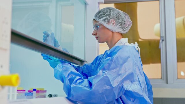 Female scientist wearing a protective suit, glasses, and hairnet working in a biosafety cabinet, mixing chemical solutions in a flask during a medical research experiment in a laboratory