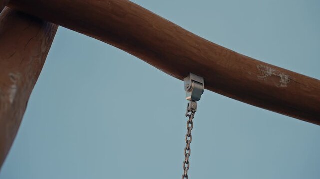 Wooden beam with metal bracket and chain sways gently against clear blue sky. natural wood grain visible knot hole in timber and metallic sheen of hardware are consistently captured