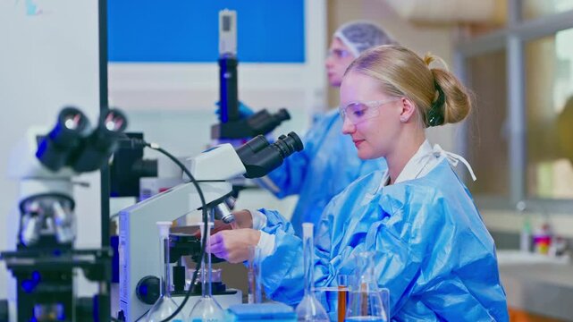 Young female scientist in a sterile laboratory conducting research using a microscope, carefully adjusting the focus to analyze samples for a new medical breakthrough or clinical study