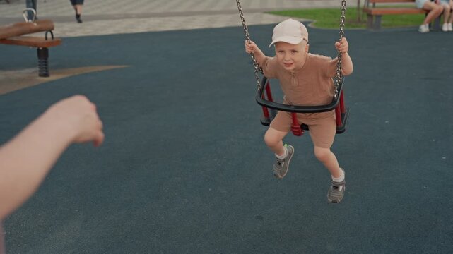 Young boy enjoys swinging at playground while adult pushes swing. scene takes place outdoors on bright daylight with other children and park equipment visible in background creating lively